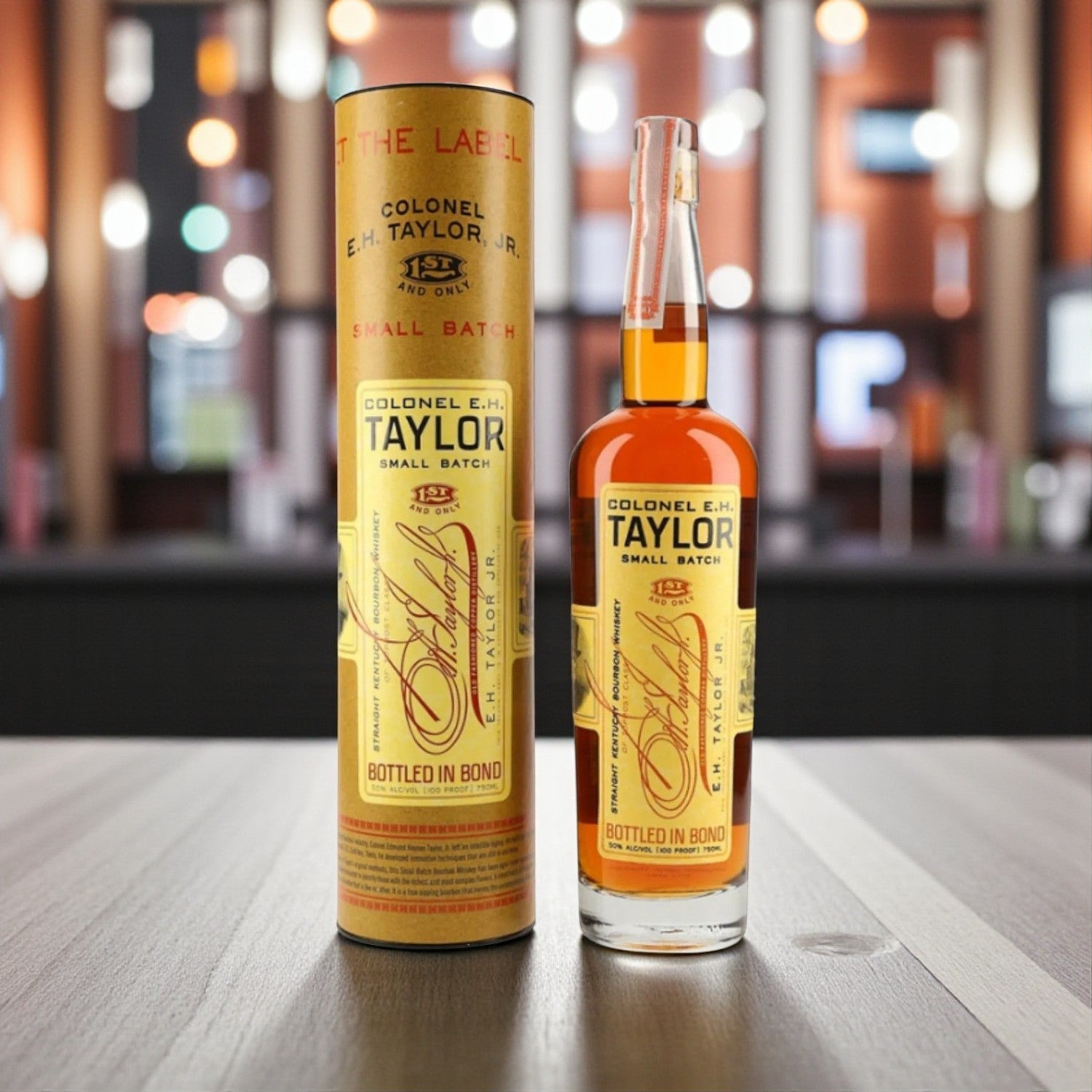 Colonel E.H. Taylor Small Batch Bottled in Bond Bourbon bottle and tube on wooden table with bar background – lifestyle shot from Total Bourbon