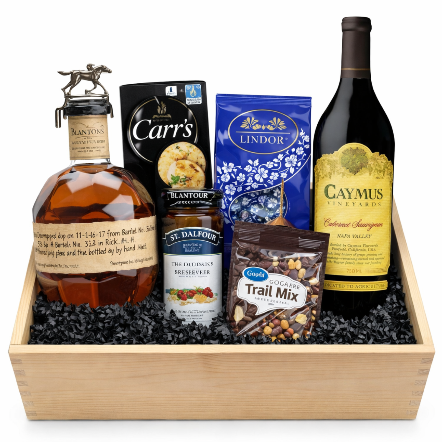 Wooden gift crate with Blanton’s bourbon, Caymus wine, Carr’s crackers, blue Lindt Lindor dark chocolate truffles, St. Dalfour strawberry preserves, and trail mix arranged on black crinkle paper against a white background.