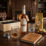 Basil Hayden's Kentucky Straight Bourbon Whiskey gift crate lifestyle scene with bottle featuring cream label and copper belt strap, old-fashioned crystal-cut whiskey glasses with neat pour, whiskey stones, Brilliant Ideas leather bourbon journal with wooden pen, Ferrero Rocher 8-piece gold bag, and open wooden crate on dark walnut table in upscale lounge setting with warm ambient lighting.