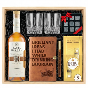 Basil Hayden's Kentucky Straight Bourbon Whiskey gift crate top-down view featuring 750ml bottle with cream label and copper belt strap, two old-fashioned crystal-cut whiskey glasses, Royal Reserve Whiskey Stones set, Brilliant Ideas leather bourbon journal with wooden pen, and Ferrero Rocher 8-piece gold bag in wooden crate with black crinkle paper on white background.