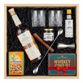 Basil Hayden's Old-Fashioned cocktail gift crate with Basil Hayden's Kentucky Straight Bourbon Whiskey with copper band and cork cap, crystal-cut old fashioned glasses, Angostura bitters, premium muddler, bar spoon, jigger, Peninsula cocktail cherries, La Perruche sugar cubes, and Royal Reserve whiskey stones set all nestled in black zigzag crinkle paper inside wooden crate on white background