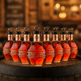 Blanton's Gold Edition Single Barrel Bourbon complete 8-bottle collector's set with gold horse and jockey stoppers and rose gold neck bands, 750ml bottles arranged in row on rustic aged oak barrel with warm amber bourbon bar lighting and moody bokeh