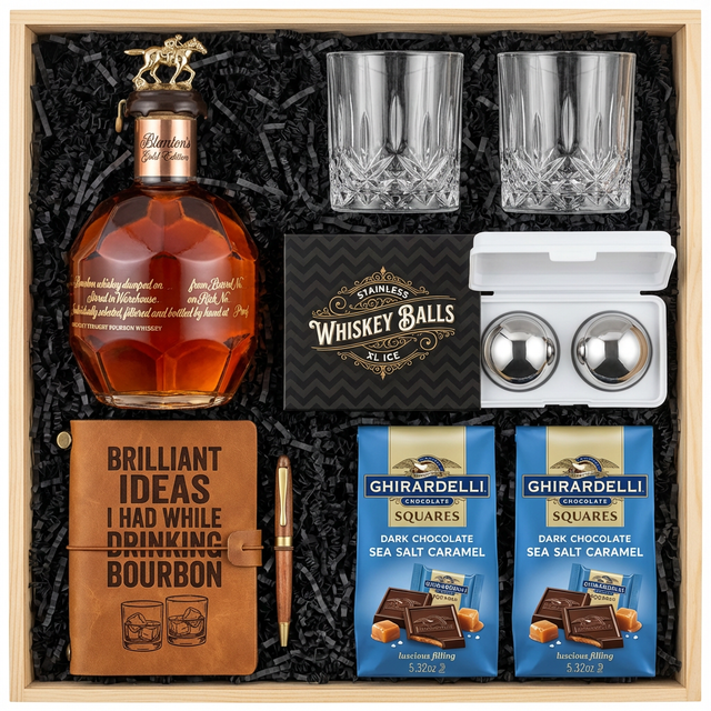 Blanton's Gold Edition Single Barrel Bourbon gift crate with gold horse stopper, crystal-cut old fashioned glasses, faux-leather bourbon journal and pen set, premium XXL stainless steel whiskey balls, and two Ghirardelli Dark Chocolate Sea Salt Caramel bags on rustic aged oak surface with warm amber bourbon bar lighting and moody bokeh
