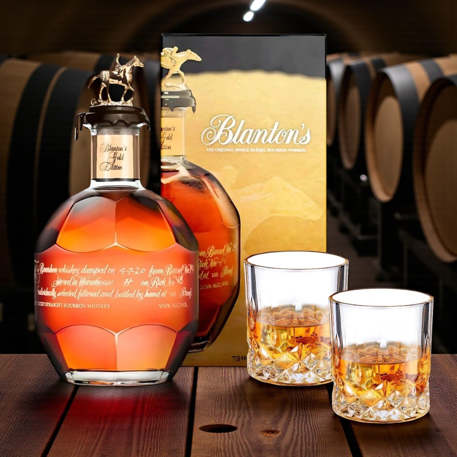 Blanton’s Gold Edition Single Barrel Bourbon Gift Set with bottle, box, and two crystal glasses on a wooden table in a barrel room – premium bourbon from Total Bourbon