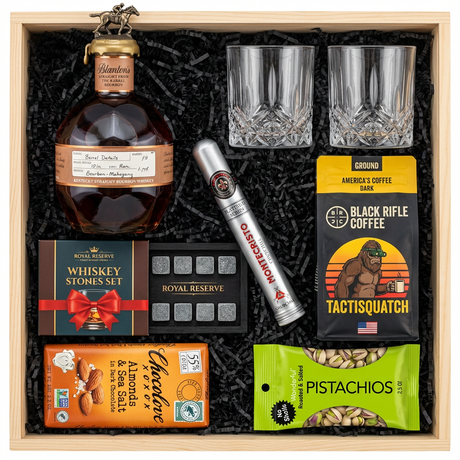 Blanton's Straight From The Barrel Single Barrel Bourbon 700ml gift crate with bronze horse stopper and gold wax cap, crystal-cut old fashioned glasses, Royal Reserve whiskey stones set with tray, Montecristo Platinum cigar tube, Black Rifle Coffee Tactisquatch blend, Chocolove Almonds & Sea Salt dark chocolate bar, and Wonderful Pistachios in black crinkle paper on white background