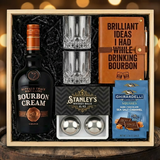 Buffalo Trace Bourbon Cream Liqueur gift crate with black bottle, crystal glasses, bourbon journal and pen set, Stanley's XXL whiskey balls, and two Ghirardelli Dark Chocolate Sea Salt Caramel bags nestled in black zigzag crinkle paper inside wooden crate on rustic wood surface with warm ambient lighting