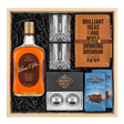 Bird's eye view of Elmer T. Lee Single Barrel Sour Mash Kentucky Straight Bourbon Whiskey gift crate with gold cap and dark brown oval label, crystal-cut old fashioned glasses, tan leather bourbon journal and pen set, Stanley's XXL stainless steel whiskey balls with rectangular freezer tray, and two Ghirardelli Dark Chocolate Sea Salt Caramel bags all nestled in black zigzag crinkle paper inside wooden crate on white background