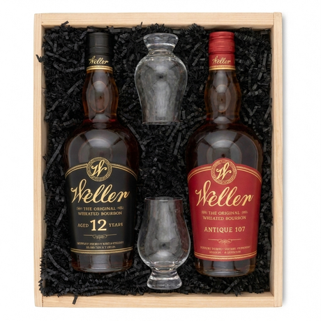 A Weller whiskey gift set in a light wood box filled with black shredded paper. The set includes two bottles of Weller bourbon whiskey, one labeled 12 Years and one labeled Antique 107, along with a small clear decanter and a small clear whiskey glass.