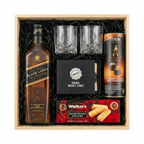 Johnnie Walker Black Label 12 Year Scotch Gift Crate featuring a 750ml bottle, two crystal cut whiskey glasses, Original Whiskey Stones set, Walker's Shortbread Fingers, and Turin Johnnie Walker Bittersweet Chocolates tube, arranged in a light wooden gift box with black crinkle paper