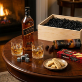 Lifestyle scene of Johnnie Walker Black Label 12 Year Scotch gift set being enjoyed on an elegant dark wood table with crystal glasses filled with scotch, whiskey stones, Walker's Shortbread Fingers, and Turin chocolates, with the wooden gift crate in the background