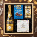 Louis Roederer Cristal Champagne gift crate with clear glass bottle and gold labels, Ghirardelli Dark Chocolate Sea Salt Caramel squares, Godiva Let's Celebrate chocolate box, and Ferrero Rocher chocolates on elegant dark wood surface with warm golden bokeh lights and celebratory decor