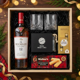 The Macallan 12 Year Sherry Oak Single Malt Scotch gift crate, crystal-cut old fashioned glasses, premium whiskey stones gift set, Ferrero Rocher chocolates, and Walker's shortbread fingers on elegant dark wood surface with warm golden bokeh lights and holiday decor