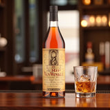 Old Rip Van Winkle 10 Year Bourbon bottle on wooden bar beside glass of bourbon on ice with warm lighting – lifestyle shot from Total Bourbon