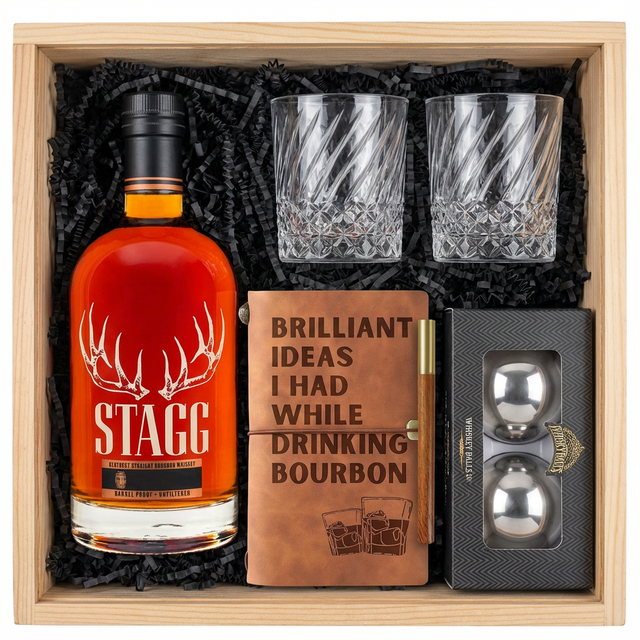 Stagg Kentucky Straight Bourbon Whiskey barrel proof gift crate with antler logo bottle, twisted crystal-cut old fashioned glasses, tan leather bourbon journal and pen set, and Stanley's XXL stainless steel whiskey balls with freezer tray all realistically nestled in black zigzag crinkle paper inside wooden crate on white background