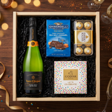 Veuve Clicquot Extra Brut Extra Old Champagne gift crate with black label and orange foil, Ghirardelli Dark Chocolate Sea Salt Caramel squares, Godiva Let's Celebrate chocolate box, and Ferrero Rocher chocolates on elegant dark wood surface with warm golden bokeh lights and celebratory decor
