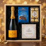 Veuve Clicquot Yellow Label Brut Champagne gift crate with Ghirardelli Dark Chocolate Sea Salt Caramel squares, Godiva Let's Celebrate chocolate box, and Ferrero Rocher chocolates on elegant dark wood surface with warm golden bokeh lights and celebratory decor